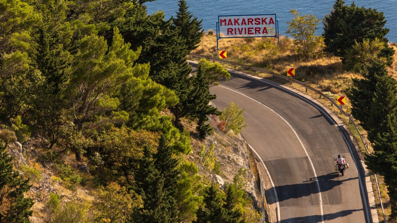 Schild zur Makarska Riviera an einer ruhigen Straße mit einer Person auf dem Fahrrad, die nach links abbiegt und an den Bäumen auf einem kleinen Hügel vorbeifährt.
